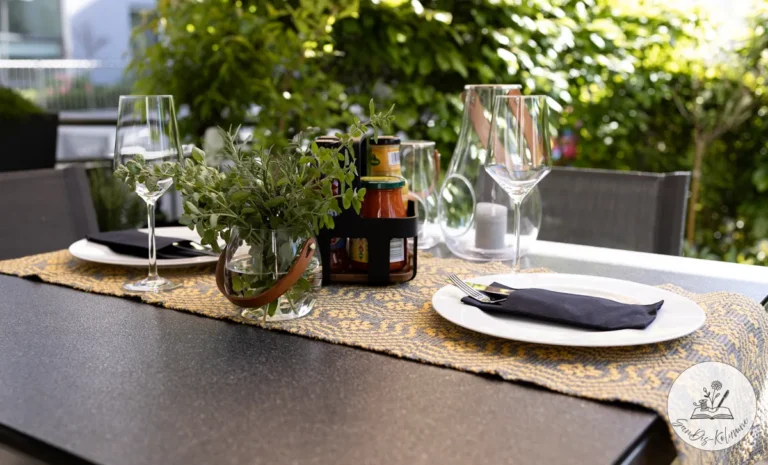 Dunkler Gartentisch mit gelb-grau gewebtem Läufer, weißen Tellern, Weingläsern, Kräuterstrauß und Windlicht vor grüner Hecke