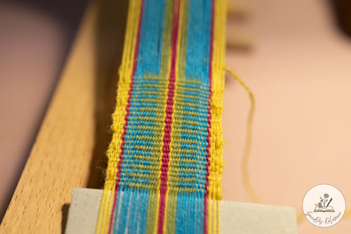 Die ersten Zentimeter Webband in Gelb, Türkis, Pink und Grün sind fertig, unten steckt ein grauer Kartonstreifen in der Kette