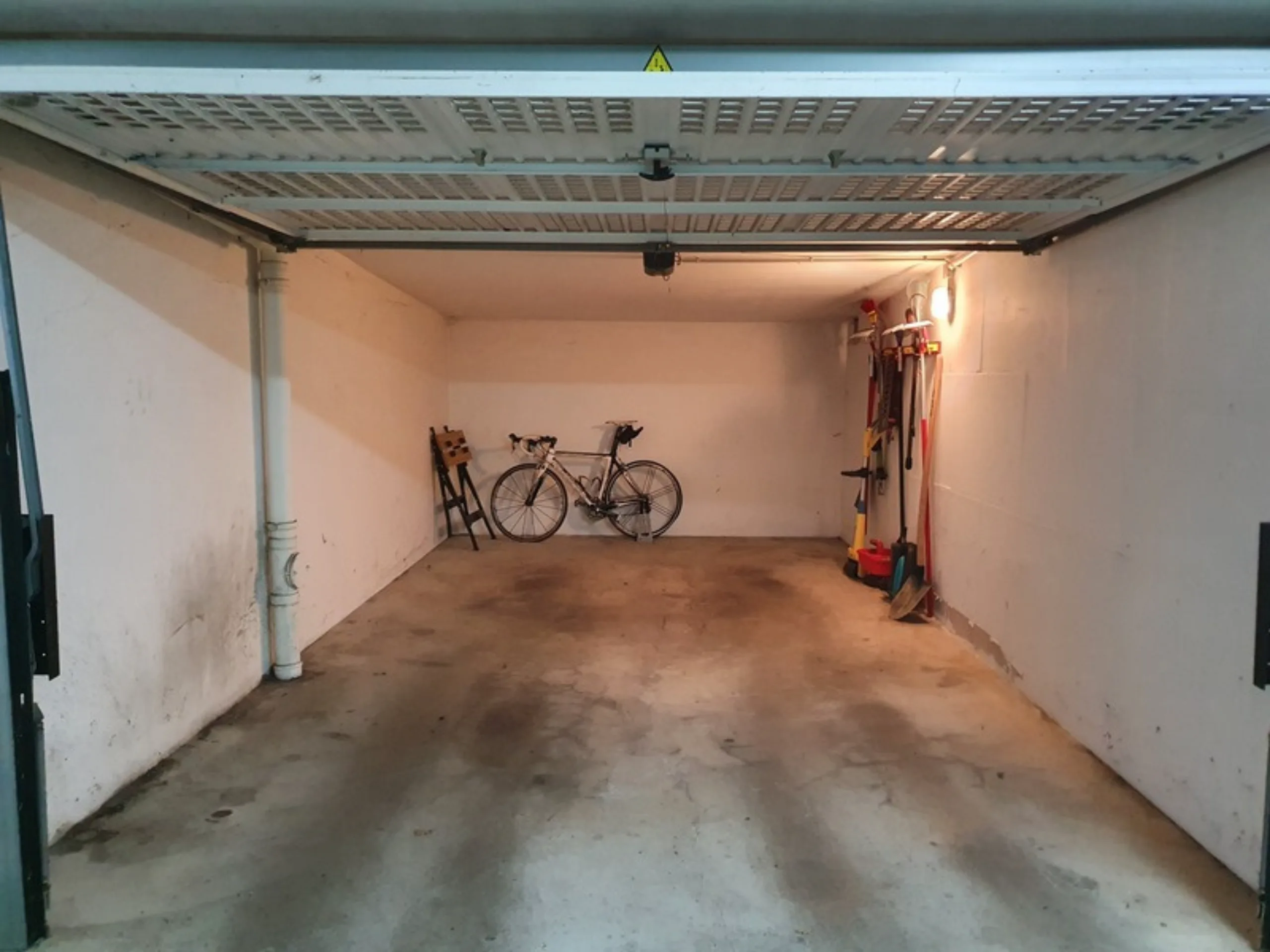 Leergeräumte Garage mit grauem Betonboden, an der Rückwand lehnt ein Rennrad, rechts hängen Besen und Gartenwerkzeuge an der Wand