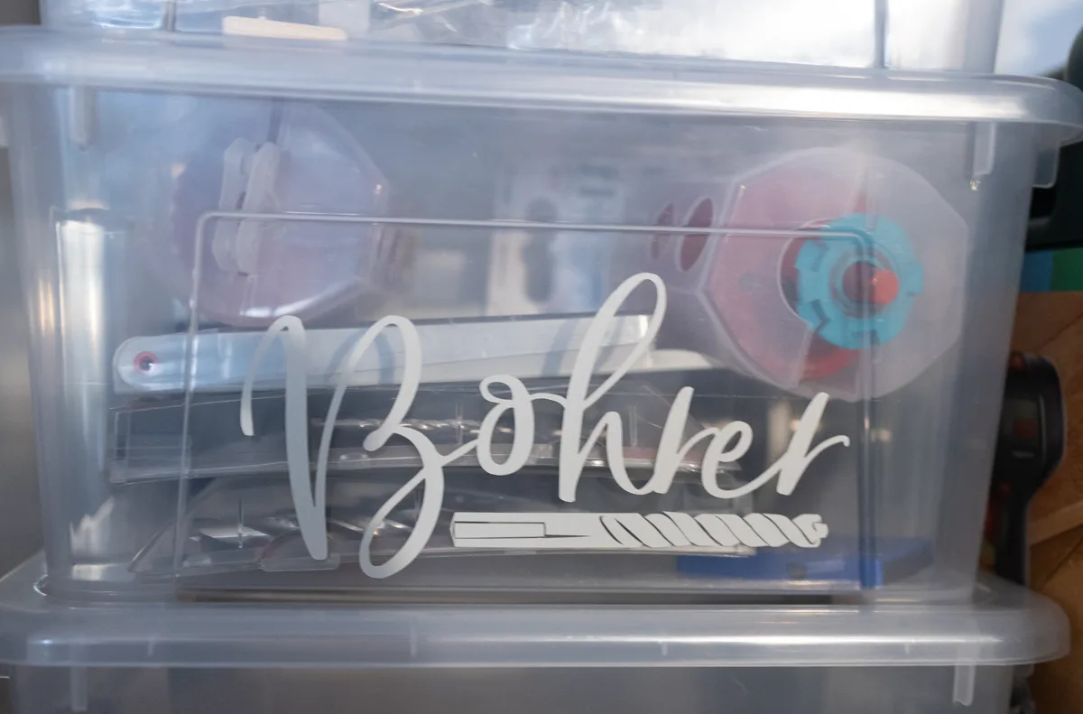 Transparente Aufbewahrungsbox mit der Aufschrift Bohrer in eleganter weißer Kalligrafie samt kleiner Bohrer-Illustration, durchs Plastik schimmert das einsortierte Werkzeug.