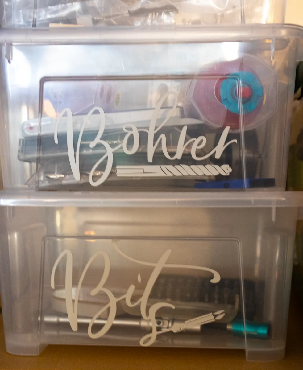 Zwei gestapelte transparente Boxen mit den Kalligrafie-Aufschriften Bohrer und Bits, gefüllt mit ordentlich sortiertem Werkzeug.