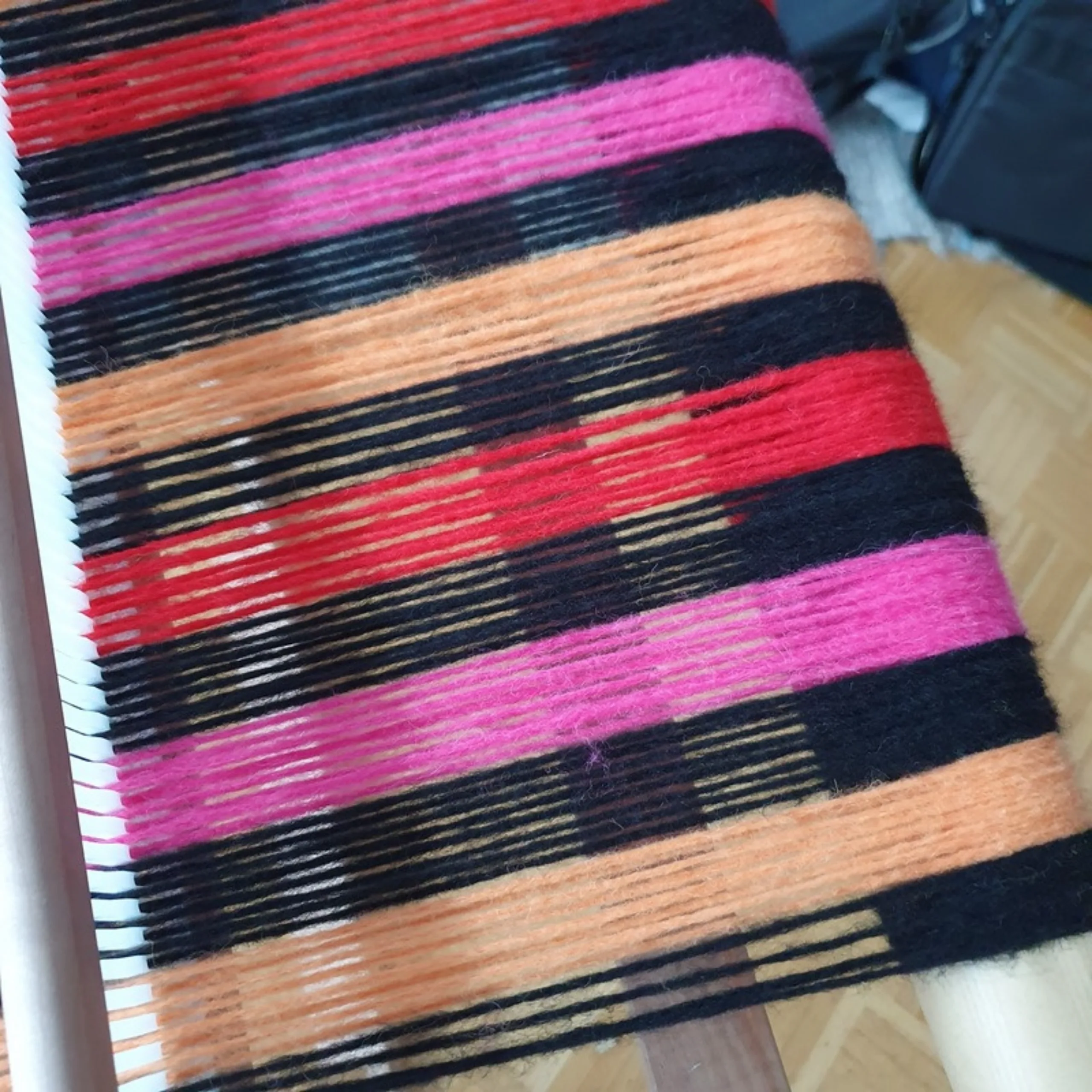 Blick über die dicht gespannten Kettfäden des Stern-Schals, die Streifen in Rot, Pink, Orange und Schwarz liegen nebeneinander