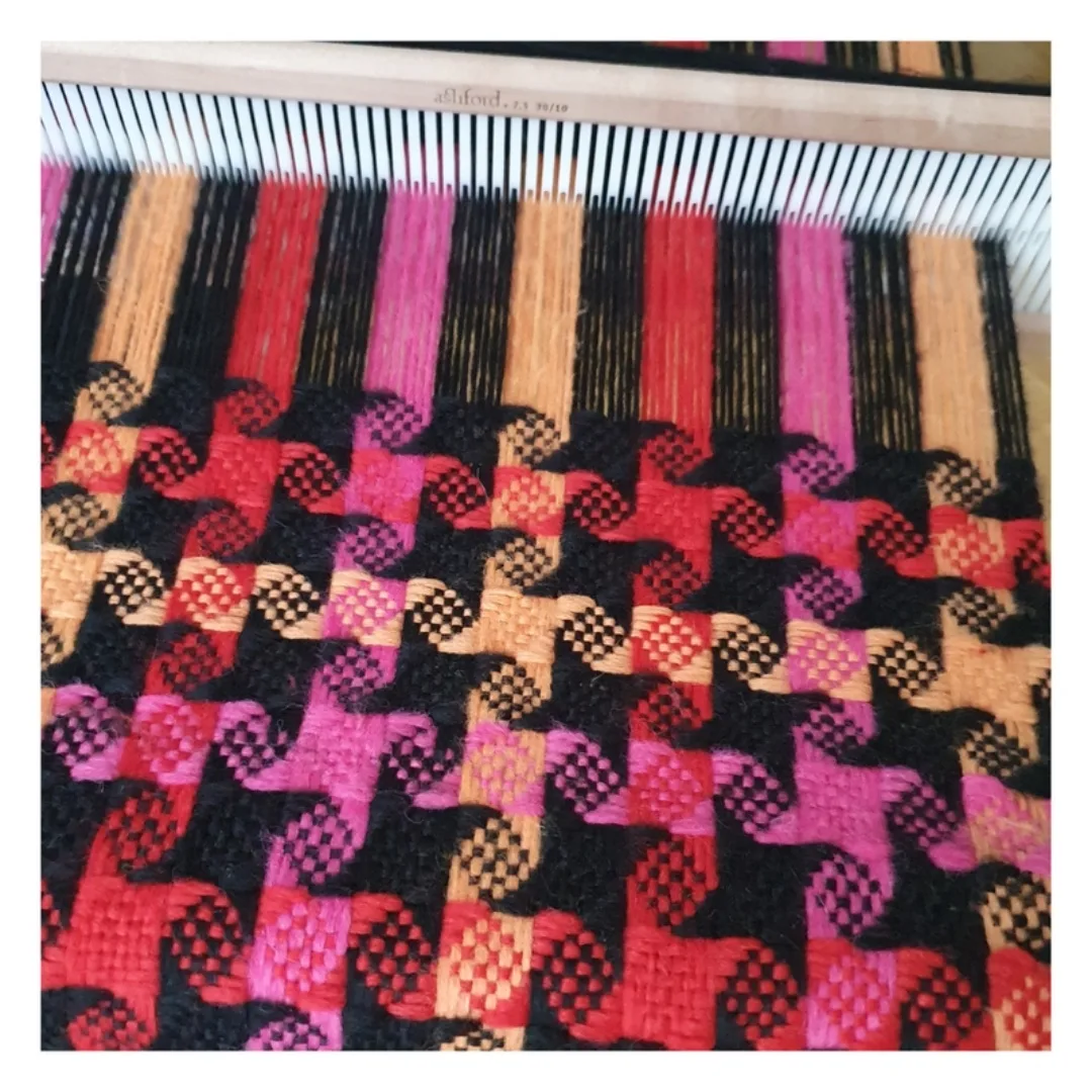 Fertig gewebtes Stück des Stern-Schals mit kleinen Windrad-Mustern in Pink, Rot, Orange und Schwarz, oben der Kamm des Webrahmens