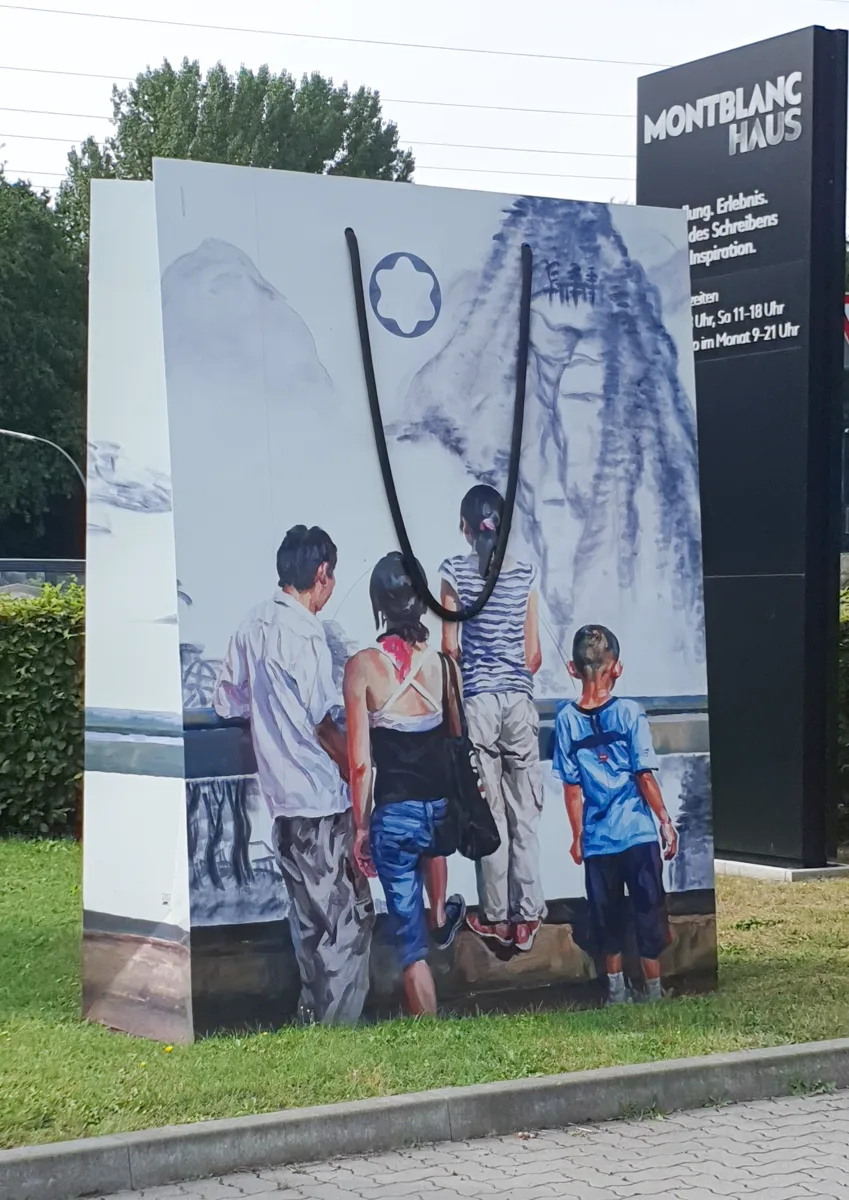 Hochformatige Taschen-Skulptur mit gemalter Familie vor Berglandschaft im Tuschestil, daneben die schwarze Eingangs-Stele des Montblanc Haus.