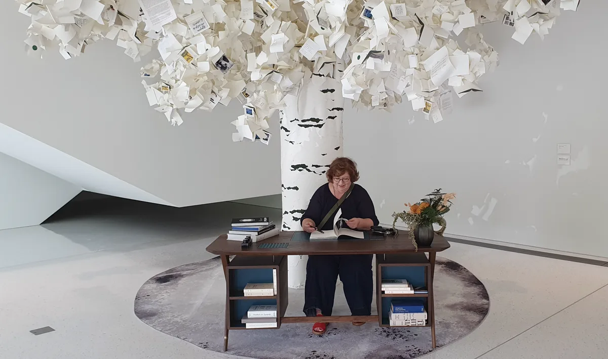 Sandra Dirks sitzt konzentriert schreibend am dunklen Holzschreibtisch unter dem Papierbaum, umgeben von Bücherstapeln und orangen Blüten.