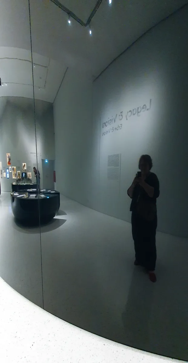 Im abgedunkelten Ausstellungsraum fotografiert eine Besucherin die leuchtende Schrift Legacy and Vision, links spiegeln sich Vitrinen in einer Glaswand.