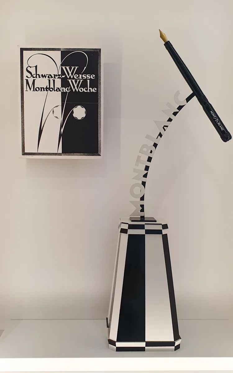 Schwarz-weißes Art-déco-Plakat der Schwarz-Weissen Montblanc Woche neben einer Skulptur mit überdimensionaler Schreibfeder auf kariertem Sockel.