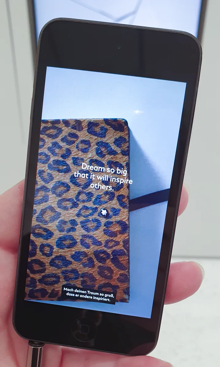 Eine Hand hält ein Smartphone mit dem Foto eines Montblanc-Notizbuchs im Leoparden-Look, überlagert vom Zitat Mach deinen Traum so groß, dass er andere inspiriert.