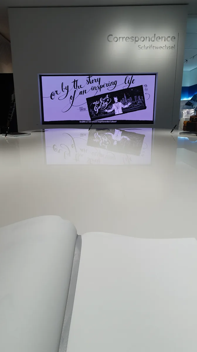 Im Raum Schriftwechsel zeigt ein großer Bildschirm Kalligrafie zu The Great Gatsby, davor ein aufgeschlagenes weißes Buch auf spiegelnder Fläche.