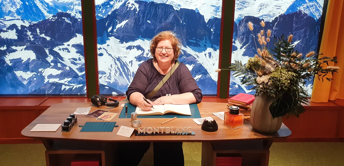 Sandra Dirks sitzt lächelnd am Filmset-Schreibtisch vor dem Mont-Blanc-Panorama und schreibt mit dem Füller in ein aufgeschlagenes Buch.