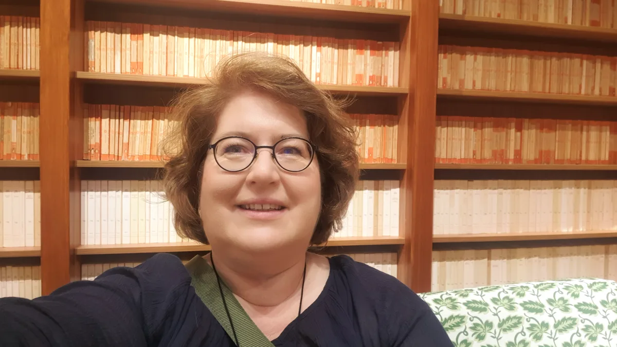 Sandra Dirks lächelt für ein Selfie vor der deckenhohen Bibliothekswand mit einheitlich gebundenen Büchern in Orange- und Beigetönen.
